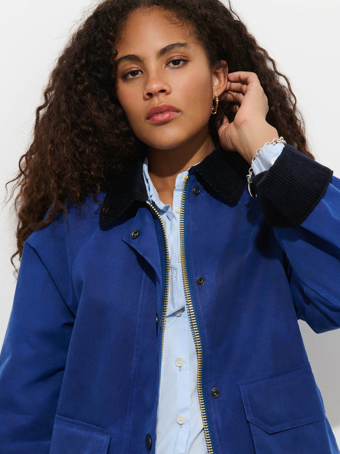A woman with curly hair wears the Chiltern Cropped Jacket In Waxed Canvas, featuring a stylish blue design with a black collar and layered over a light blue shirt. She touches her hair and looks calmly at the camera against a plain background, effortlessly showcasing modern women’s outerwear.