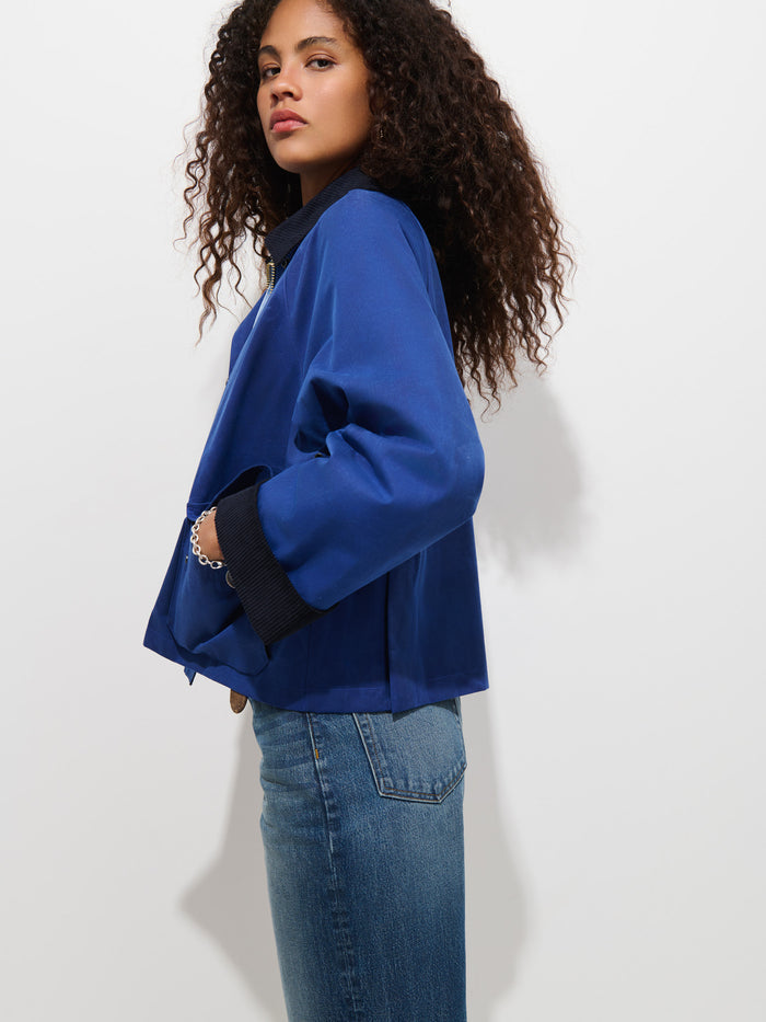 A woman with curly hair models the Chiltern Cropped Jacket In Waxed Canvas in blue, paired with jeans and standing sideways with one hand in her pocket against a plain white background.