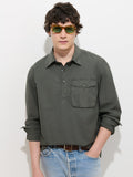 A young man with wavy dark hair and sunglasses stands against a plain white background with his hands in his pockets, wearing the Clint Popover in Cotton Poplin—an olive-green garment-dyed men’s shirt with a chest pocket—paired with light blue jeans.