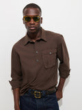 A man wearing yellow sunglasses, the Clint Popover in Cotton Poplin—a garment-dyed brown button-up men’s shirt with a chest pocket—and dark jeans stands against a plain white background with his hands in his pockets.