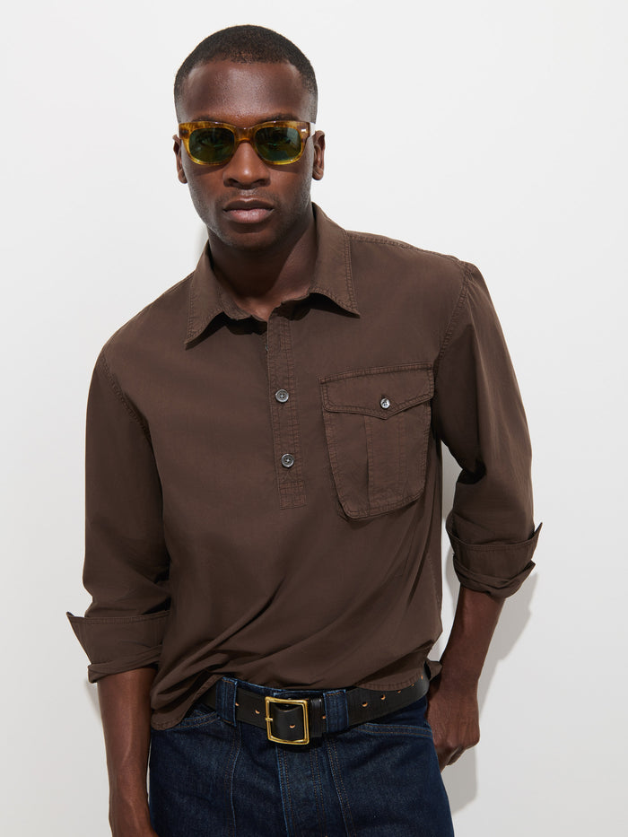 A man wearing yellow sunglasses, the Clint Popover in Cotton Poplin—a garment-dyed brown button-up men’s shirt with a chest pocket—and dark jeans stands against a plain white background with his hands in his pockets.