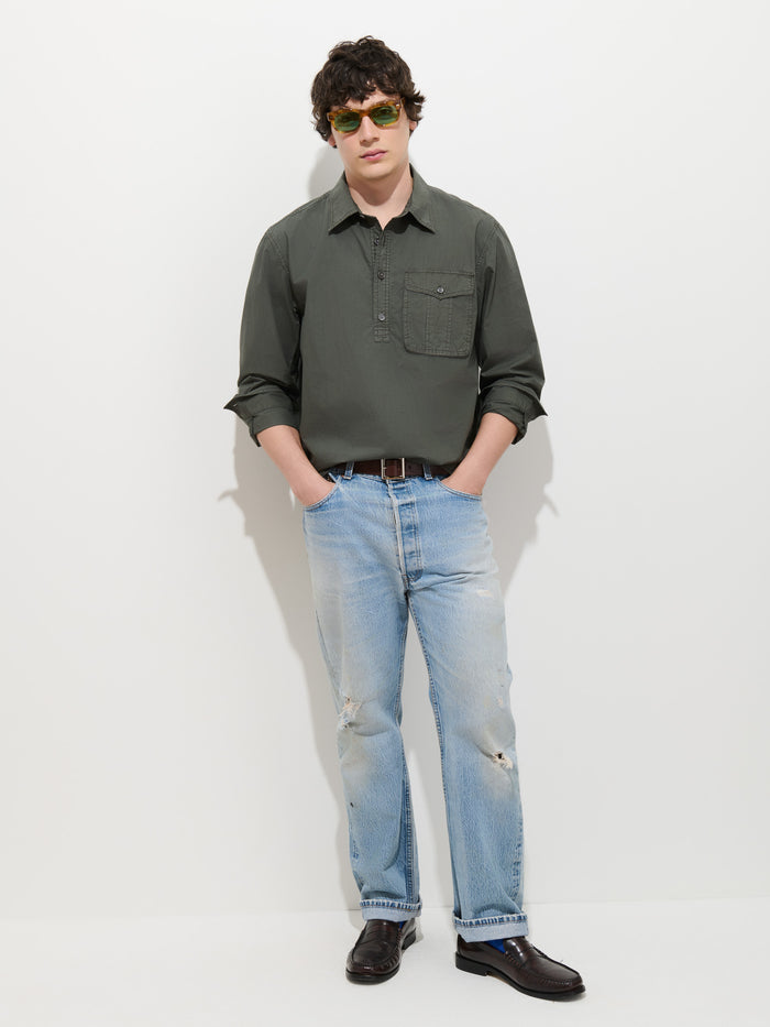 A person wearing green sunglasses, the Clint Popover in Cotton Poplin—a dark green garment-dyed men’s shirt—light blue distressed jeans, and dark loafers stands against a plain white background with hands in pockets.