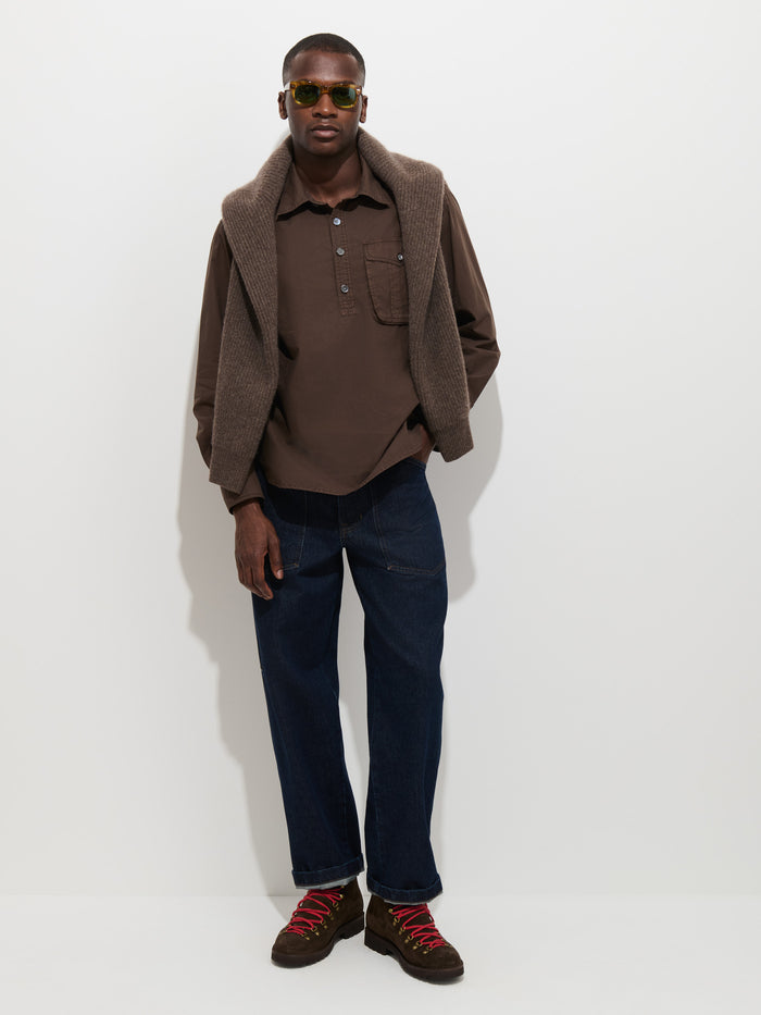 Man stands against a white wall wearing sunglasses, the Clint Popover in Cotton Poplin—a garment-dyed brown button-up men’s shirt—paired with a brown sweater draped over his shoulders, blue jeans, and brown shoes with red laces.