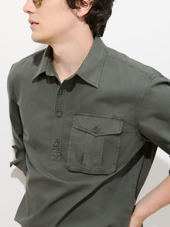 A person wearing sunglasses and the Clint Popover in Cotton Poplin—a green men’s shirt made from garment-dyed cotton with a front pocket—stands against a plain white background.