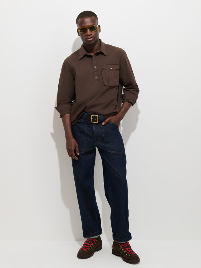 A man wearing sunglasses, the Clint Popover in Cotton Poplin—a brown garment-dyed long-sleeve polo shirt—blue jeans, and brown hiking boots with red laces stands against a plain white background.