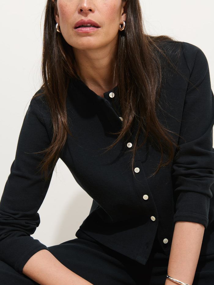 A woman with long brown hair wears the Coco Sport Cardigan, featuring troca shell buttons, paired with silver hoop earrings. She is seated with one arm on her leg, facing slightly to the side against a plain light background.