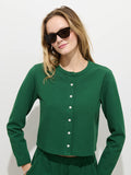 Wearing the Coco Sport Cardigan in emerald stretch cotton jersey with Troca shell buttons, a woman poses against a white background. Her loose light brown hair and black sunglasses complete her chic, stylish look and slight smile.