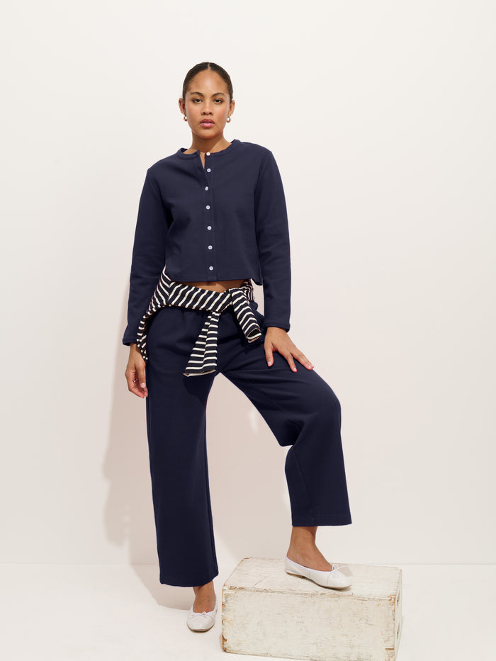 A woman stands against a plain light background, wearing a navy stretch cotton jersey button-up top and matching Coco Sport Cardigan pants. A black-and-white striped sweater is tied at her waist, and she wears white flats with one foot on a white wooden box.