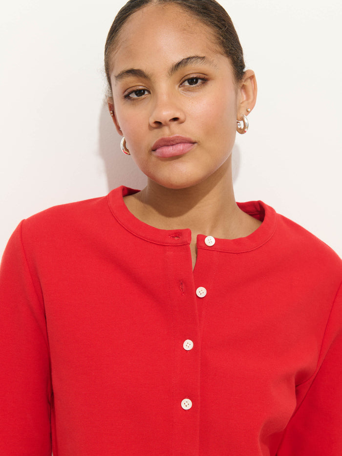 A woman with pulled-back hair confidently models the Coco Sport Cardigan, a bright red stretch cotton jersey top with white buttons, paired with small hoop earrings against a plain white background, showcasing chic designer style.