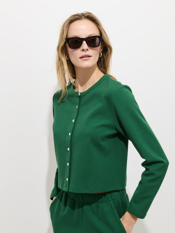 A woman in black sunglasses and the Coco Sport Cardigan—an emerald stretch cotton jersey women’s designer set with Troca shell buttons—stands against a plain white background, looking confidently at the camera.
