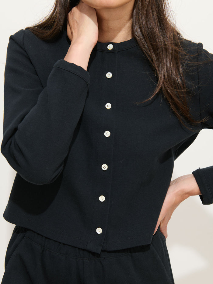 A woman models the Coco Sport Cardigan—a long-sleeve black stretch cotton jersey top with white troca shell buttons—standing with one hand on her hip and the other at her neck; her face is partially out of frame.