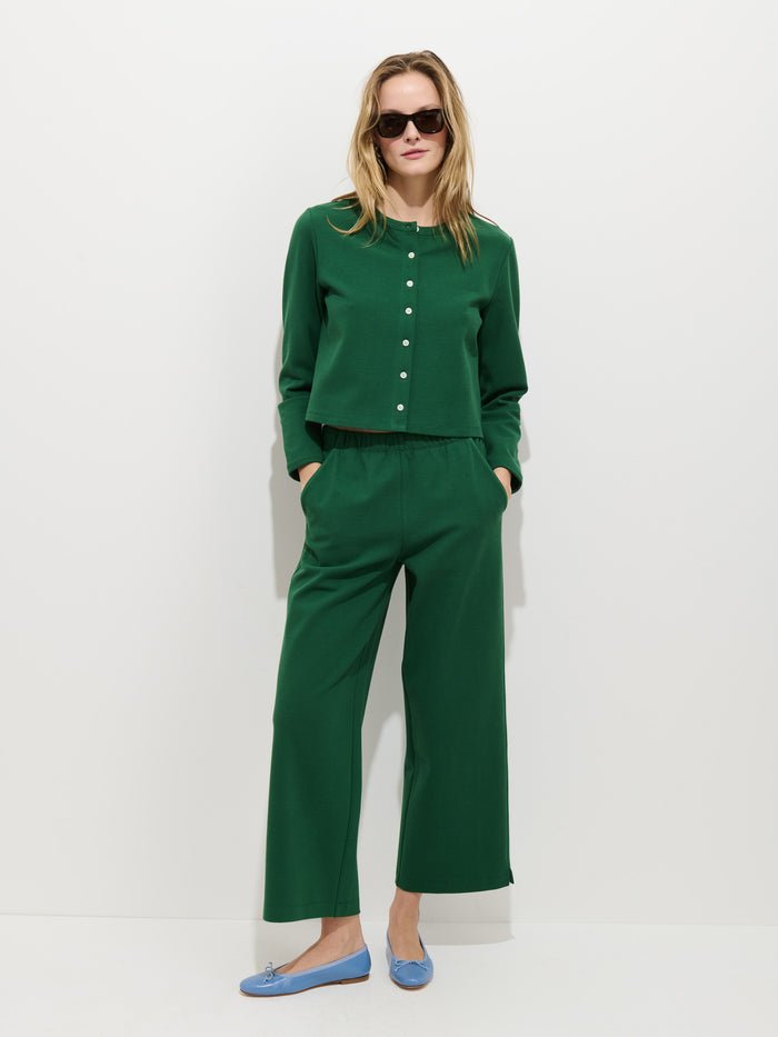 A person wears the Coco Sport Cardigan, an emerald stretch cotton jersey women’s designer set with Troca shell buttons. They stand against a white background with hands in pockets, sunglasses, and light blue flats.