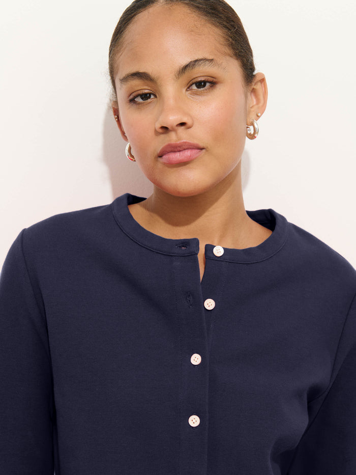 A woman with medium skin tone and short, pulled-back hair wears silver hoop earrings, a navy button-up shirt, and the Coco Sport Cardigan in stretch cotton jersey, looking calmly at the camera against a plain light background.