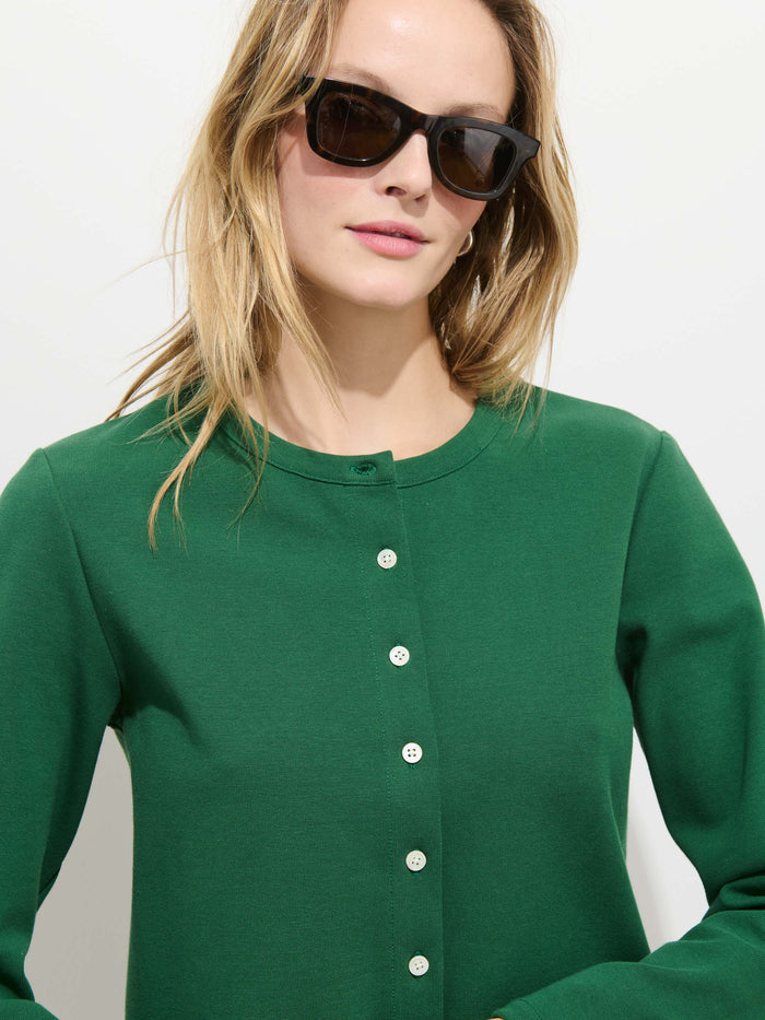 A woman with long blonde hair and black sunglasses wears the Coco Sport Cardigan—an emerald stretch cotton jersey with Troca shell buttons—standing against a plain white background.