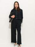 A woman stands against a white wall in Coco Sport Pants and a black stretch cotton jersey top with pearl buttons, silver flats, and a textured black clutch—a chic designer ensemble.