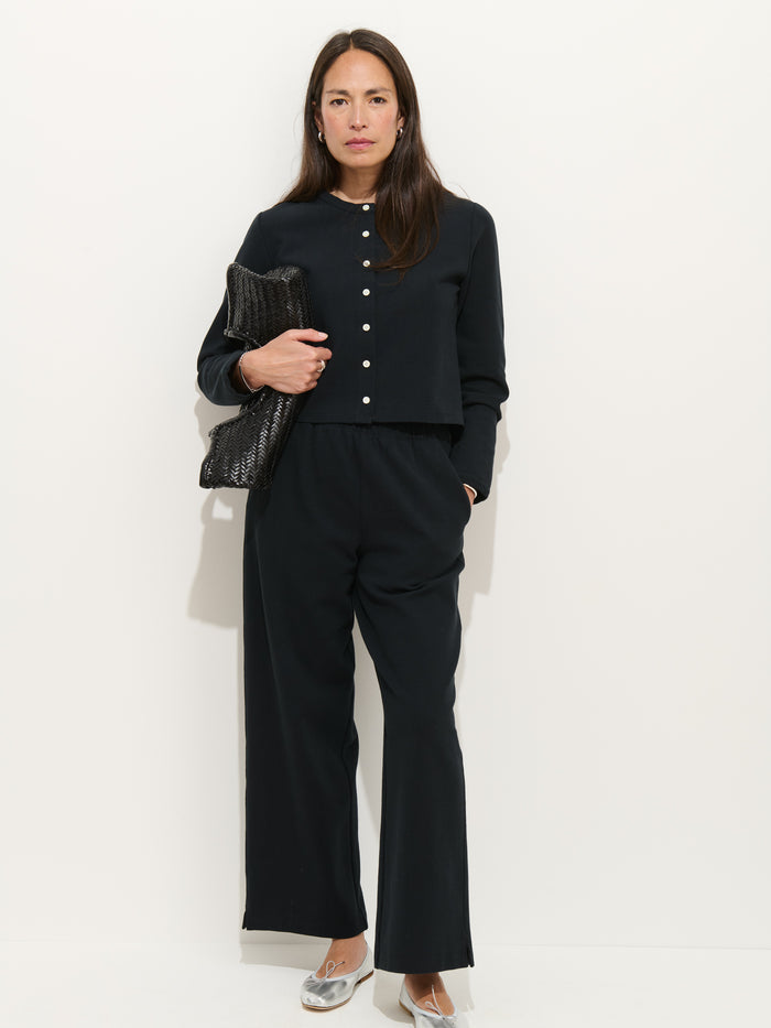 A woman stands against a white wall in Coco Sport Pants and a black stretch cotton jersey top with pearl buttons, silver flats, and a textured black clutch—a chic designer ensemble.