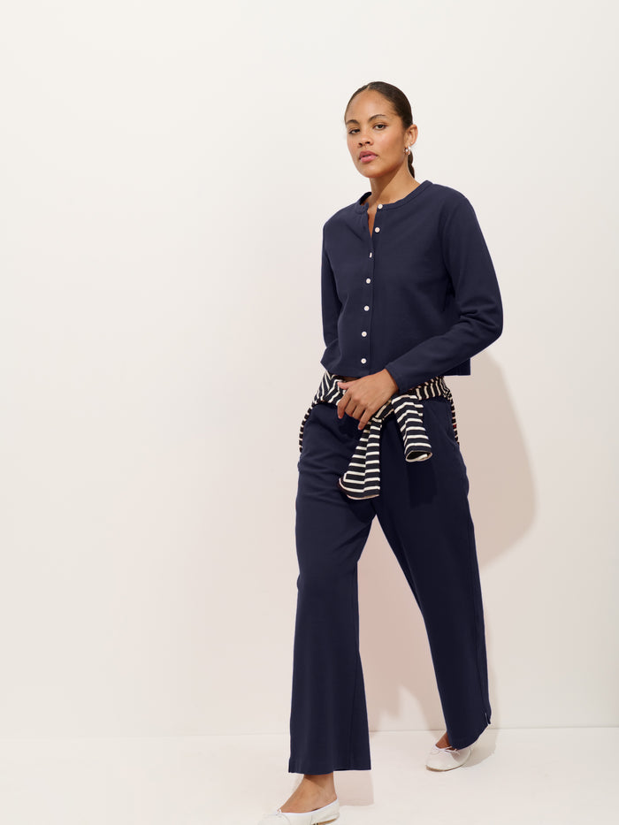 A woman in a navy long-sleeve button-up and Coco Sport Pant in stretch cotton jersey walks forward. A striped sweater is tied at her waist; she wears white shoes against a plain light background, showcasing effortless women’s designer style.