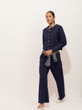 A woman in a navy long-sleeve button-up and Coco Sport Pant in stretch cotton jersey walks forward. A striped sweater is tied at her waist; she wears white shoes against a plain light background, showcasing effortless women’s designer style.