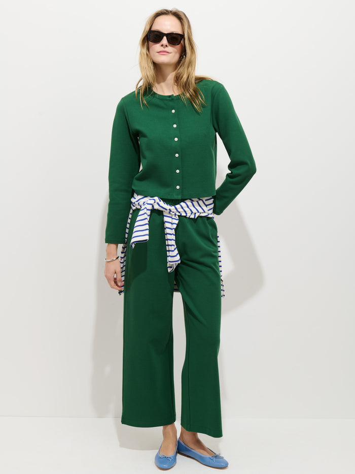 A woman stands against a white background wearing a green buttoned stretch cotton jersey top and matching Coco Sport Pant. She accessorizes with sunglasses, blue flats, and a striped shirt tied around her waist for a chic look.