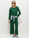 A woman stands against a white background wearing a green buttoned stretch cotton jersey top and matching Coco Sport Pant. She accessorizes with sunglasses, blue flats, and a striped shirt tied around her waist for a chic look.