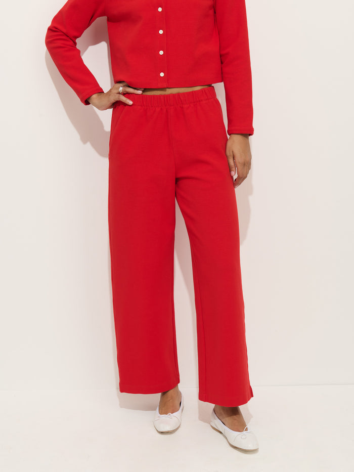 A person stands against a white background, wearing the Coco Sport Pant and a matching red long-sleeve top. Only their body from shoulders down is visible. They wear white flat shoes and have one hand on their hip.