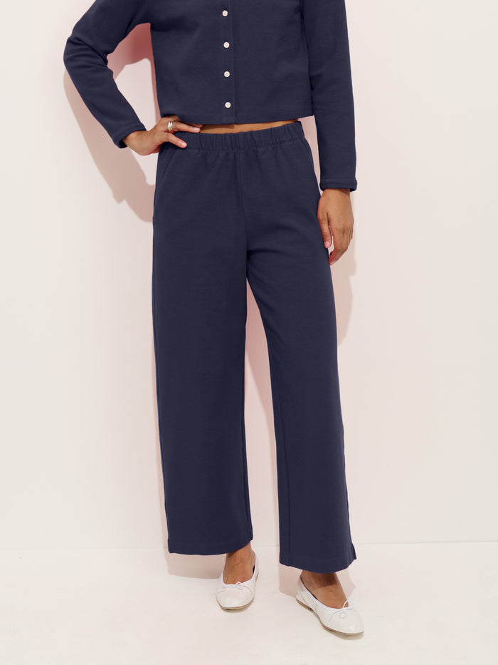 A person wears a navy blue women’s designer long-sleeve button-up top and matching Coco Sport Pant in stretch cotton jersey with white flat shoes, standing against a light background. Only the body from shoulders down is visible.