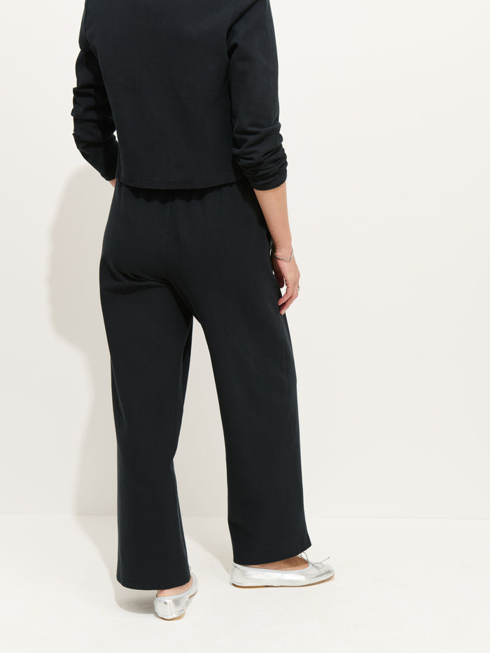 A person stands facing away, wearing a black women's designer long-sleeve top and loose Coco Sport Pants with one arm bent, white ballet flats, photographed against a plain white background.