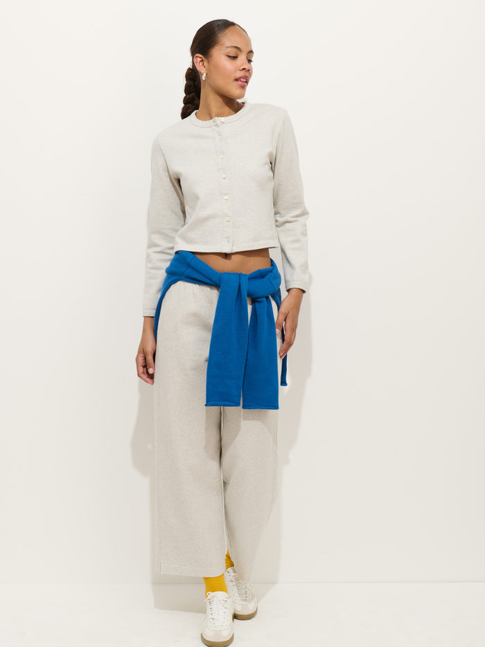 A woman stands against a white wall, wearing Coco Sport Pant and a light gray cotton double jersey button-up top. She has a blue sweatshirt tied at her waist, yellow socks, and white sneakers, looking to the side and smiling in this chic designer look.