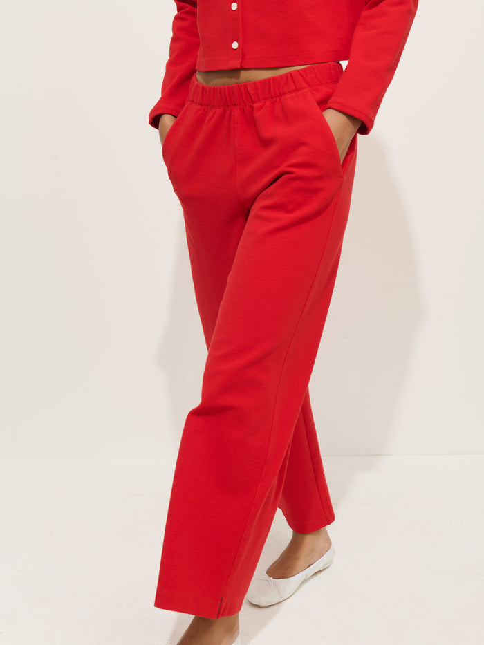 A person wears red Coco Sport Pants made from stretchy cotton jersey, standing barefoot with hands in pockets. Only their lower body is visible against a plain light background.