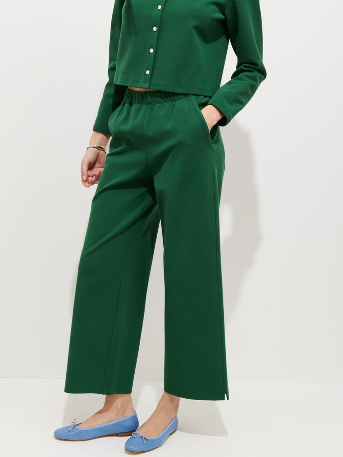 A person wears a women’s designer long-sleeve green top with buttons and matching Coco Sport Pant in stretch cotton jersey, standing with one hand in their pocket and light blue flats against a plain white background.