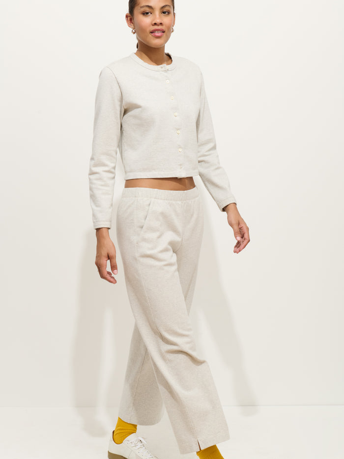 A person wears a light beige buttoned cardigan with matching Coco Sport Pants in cotton double jersey, hair pulled back, yellow socks, and white sneakers, walking confidently against a plain white background.