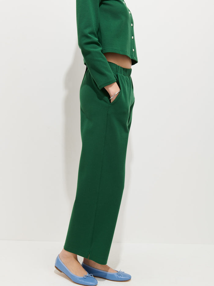 A person stands sideways wearing a green women’s designer long-sleeve top and matching Coco Sport Pant in stretch cotton jersey with hands in pockets, paired with light blue loafers, against a plain white background.