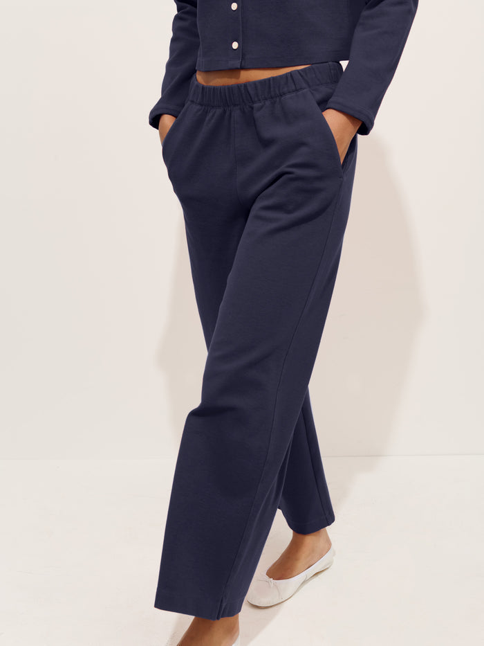 A person stands on a light floor, hands in pockets, wearing navy blue Coco Sport Pant in stretch cotton jersey with a matching long-sleeve top. White ballet flats complete this women’s designer outfit, shown from shoulders down.