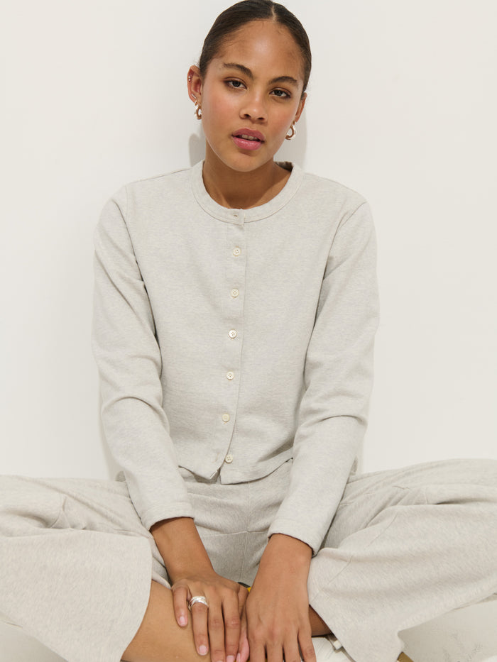 A woman wearing a light gray cotton double jersey buttoned cardigan and matching Coco Sport Pant sits cross-legged on the floor against a plain white background, looking at the camera with a neutral expression.
