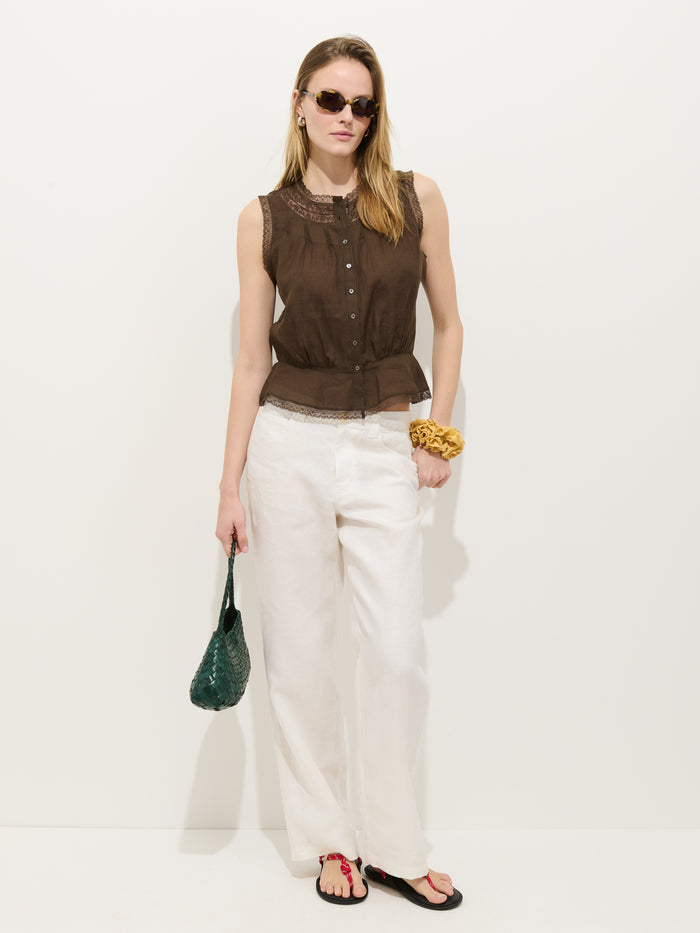 A woman in sunglasses and the Collette Top In Ramie, paired with white wide-leg pants and black sandals, stands against a white background. She carries a green woven handbag and wears a yellow scrunchie on her wrist.