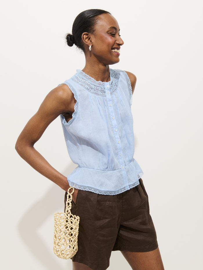 A woman smiles against a plain background, wearing the Collette Top in Ramie—a light blue, sleeveless vintage-inspired top with lace details—paired with dark brown shorts and a small woven bag. Her hair is styled in a neat bun.