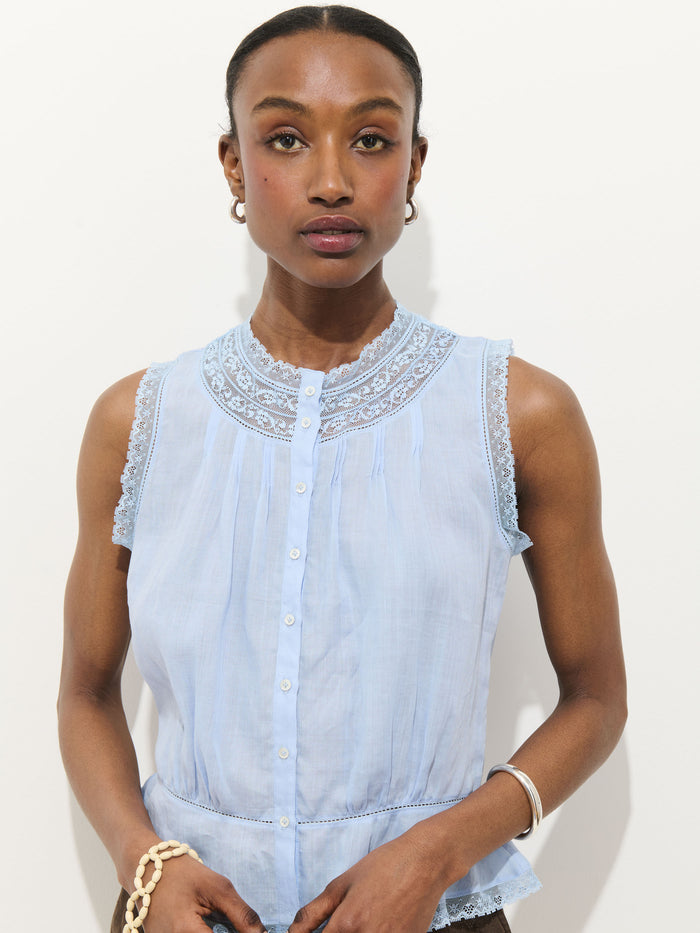 A woman with a neutral expression wears the Collette Top in Ramie, a sleeveless light blue vintage-style blouse with lace trim and button details, paired with hoop earrings and a bracelet, against a plain white background.