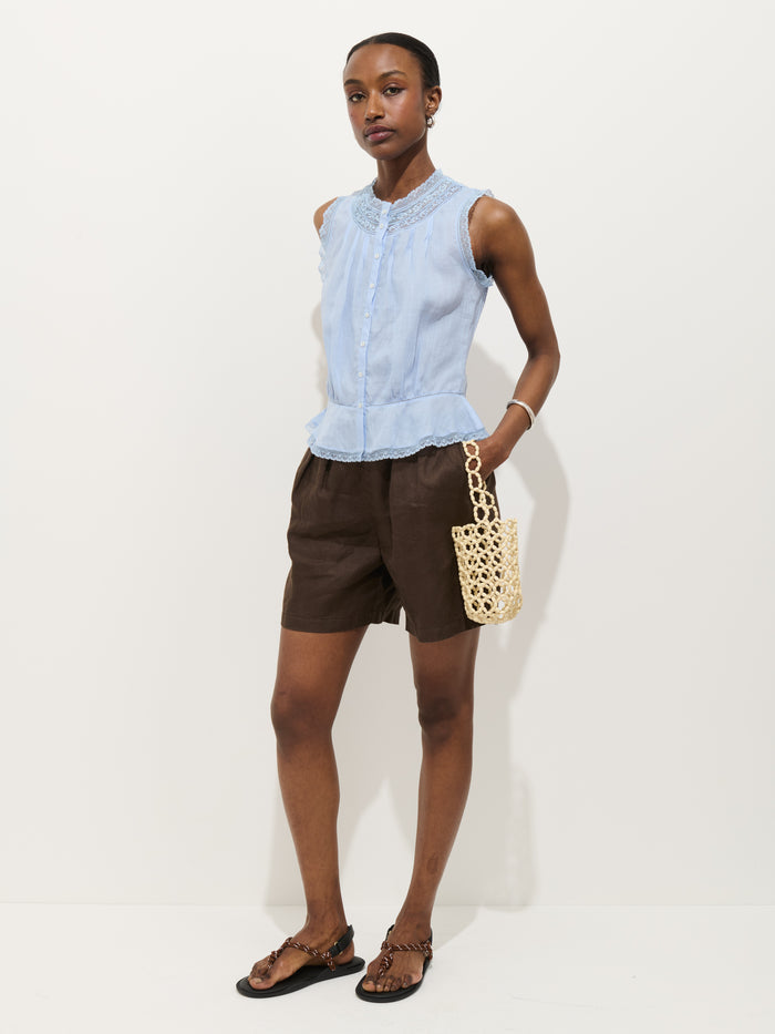 A woman stands against a white background wearing the Collette Top In Ramie, a sleeveless light blue blouse, paired with brown shorts, dark sandals, and holding a small netted cream-colored handbag.
