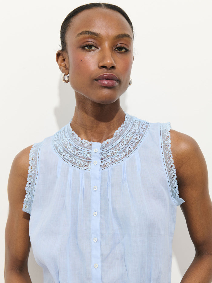 A woman models the Collette Top In Ramie, a sleeveless, light blue vintage blouse with lace trim on the collar and shoulders, looking directly at the camera against a plain white background.