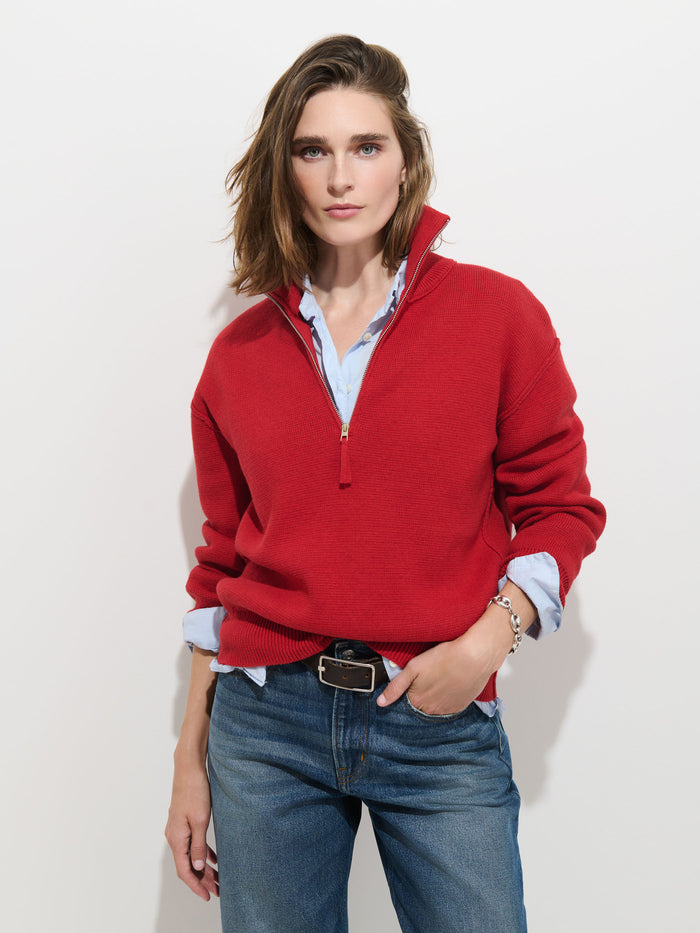 A person with shoulder-length hair wears the Daphne Half-Zip Sweater in Cotton Cashmere in red over a light blue shirt and blue jeans, standing against a plain white background with a relaxed expression.