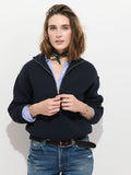 A person with shoulder-length brown hair wears the Daphne Half-Zip Sweater in Cotton Cashmere in black over a blue striped shirt, paired with blue jeans and a green scarf around their neck, standing against a white background.