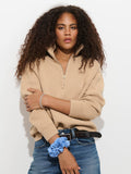 A person with curly hair wearing the Daphne Half-Zip Sweater in Cotton Cashmere—an oversized beige style—paired with blue jeans stands against a white background, arms crossed. They accessorize with a blue scrunchie and bracelet on their wrist.