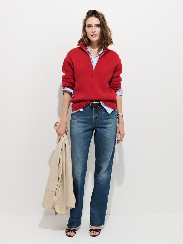 A woman with shoulder-length brown hair stands against a white background, smiling slightly as she wears the Daphne Half-Zip Sweater in Cotton Cashmere (oversized fit, red), paired with blue jeans and black sandals. She holds a beige coat in her left hand.