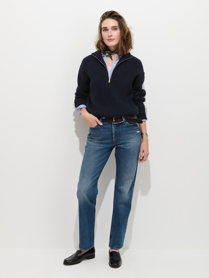 A person stands against a white background wearing the Daphne Half-Zip Sweater in Cotton Cashmere in navy, paired with blue jeans, black loafers, a striped shirt underneath, and a green neck scarf. They have one hand in their pocket and wear a relaxed expression.