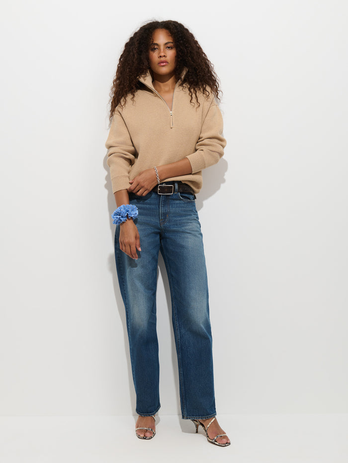 A woman with curly hair models the Daphne Half-Zip Sweater in Cotton Cashmere, styled in an oversized fit and beige color. She pairs it with blue jeans and heels, standing against a white background with one hand in her pocket and a blue scrunchie on her wrist.