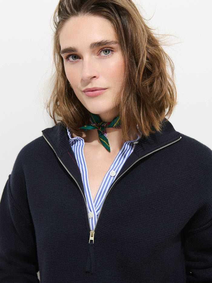 A woman with shoulder-length brown hair wears the Daphne Half-Zip Sweater in Cotton Cashmere in navy over a blue striped shirt and a green patterned neck scarf, standing against a white background.