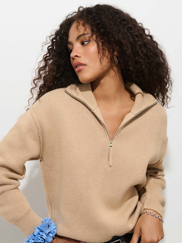 A person with curly hair is seen looking to the side, wearing the Daphne Half-Zip Sweater in Cotton Cashmere in a beige hue with an oversized fit. They have a blue scrunchie on their wrist and a chain bracelet, standing against a plain white background.