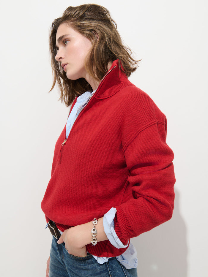 A person with shoulder-length brown hair stands against a plain white background, looking to the left, wearing the Daphne Half-Zip Sweater in Cotton Cashmere in red over a light blue shirt and blue jeans.