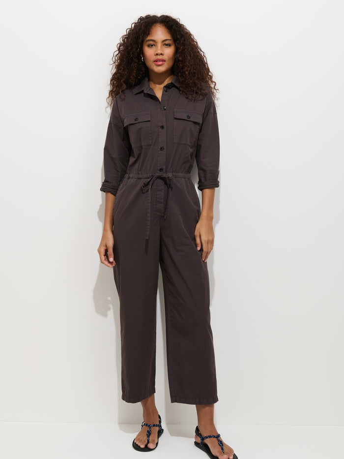 A woman with curly hair stands against a white background, wearing the Ricard Jumpsuit in Summer Twill—a dark brown, relaxed fit cotton jumpsuit with long sleeves, waist tie, and black sandals.
