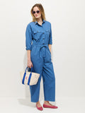 A woman in the Delancey Jumpsuit in Lightweight Twill, wearing pink sunglasses and shoes, stands against a white background holding a beige tote bag with blue handles and a red letter E.